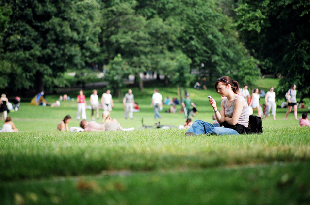 park, žena sedí na trávníku a má mobil v ruce